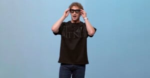 A man wearing black smart glasses, a black t-shirt, and jeans stands against a light blue background, holding the glasses with both hands and smiling.