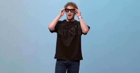 A man wearing black smart glasses, a black t-shirt, and jeans stands against a light blue background, holding the glasses with both hands and smiling.