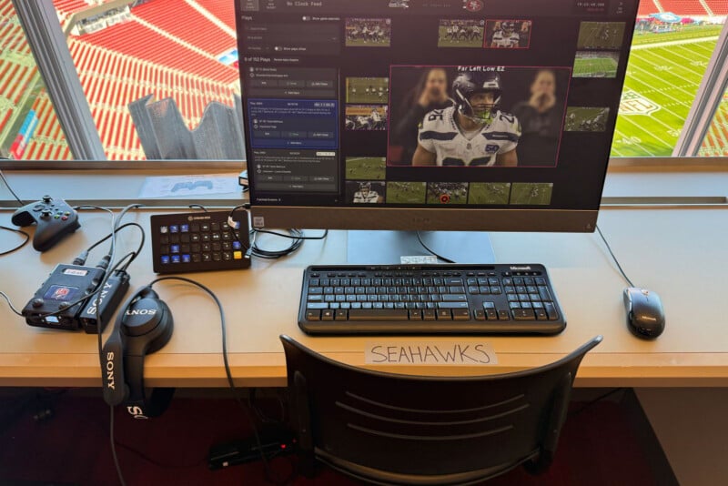 A desk with a computer monitor displaying football footage, keyboard, mouse, headphones, camera, and game controller, with "SEAHAWKS" labeled on the chair. A football stadium is visible through the window behind the desk.