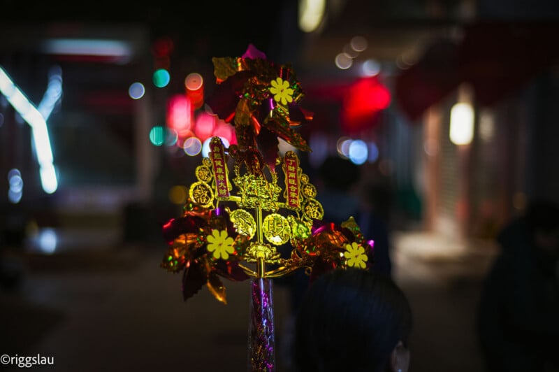 A bright, colorful decorative ornament with gold and red details is held up at night, glowing against a blurred urban background with red and yellow lights.