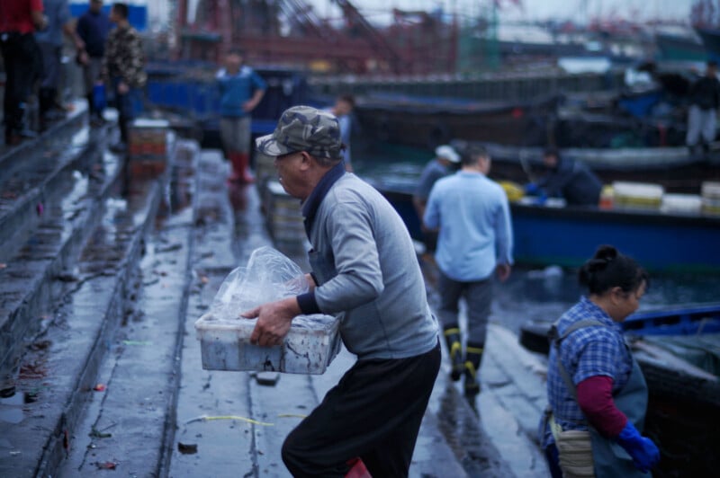 A man carries a plastic container up wet stone steps at a busy dock, surrounded by other workers and boats, with a blurred, overcast background.