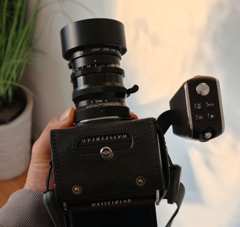 A person holds a Hasselblad medium format camera with a large lens, displaying control dials and a digital viewfinder. A green potted plant is blurred in the background.