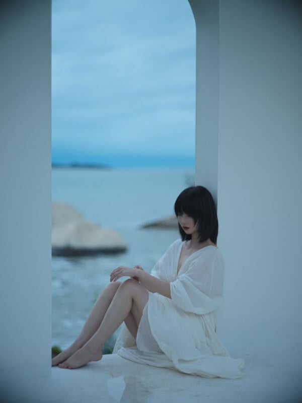 A woman in a white dress sits on a white ledge by the sea, framed by an arched doorway, with soft natural light and a calm, blue-toned atmosphere.