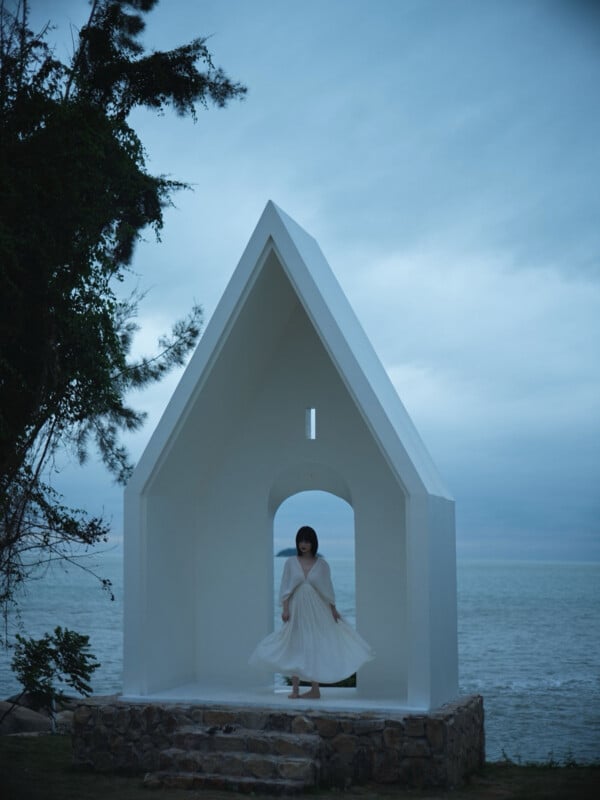 A woman in a white dress stands inside a small, white, chapel-like structure by the sea at dusk, with cloudy skies and trees nearby.
