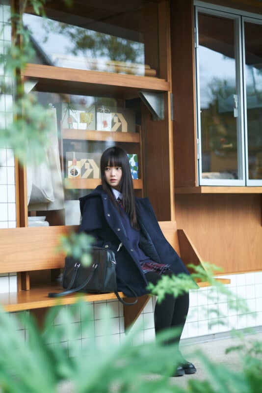 A young woman with long dark hair sits alone on a wooden bench outside a building, wearing a dark coat and holding a black bag. She gazes forward with a calm expression. Green leaves are blurred in the foreground.