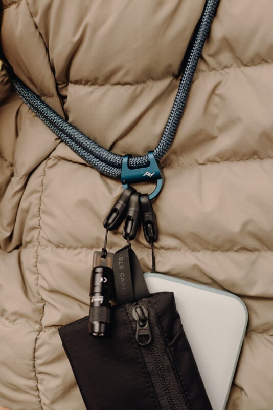Close-up of a beige puffer jacket with a blue lanyard or cord around the neck, holding a carabiner with attached items including a small flashlight, a zippered pouch, and a slim rectangular device.