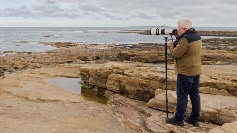 An older man stands on rocky terrain by the sea, using a camera with a large telephoto lens on a tripod, aiming toward the ocean under a cloudy sky.