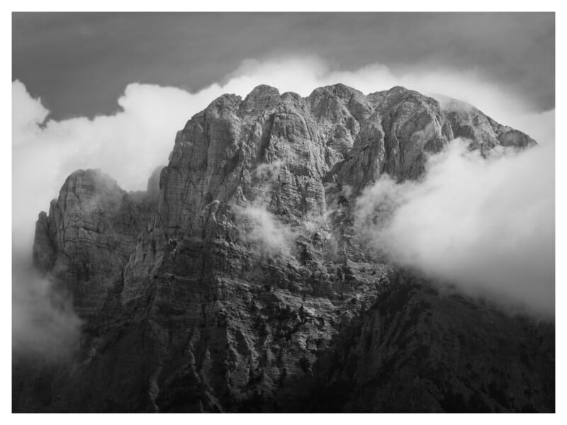 La fotografía en blanco y negro muestra un pico rocoso parcialmente cubierto de nubes, con sombras dramáticas y texturas rugosas visibles en la superficie de la roca.