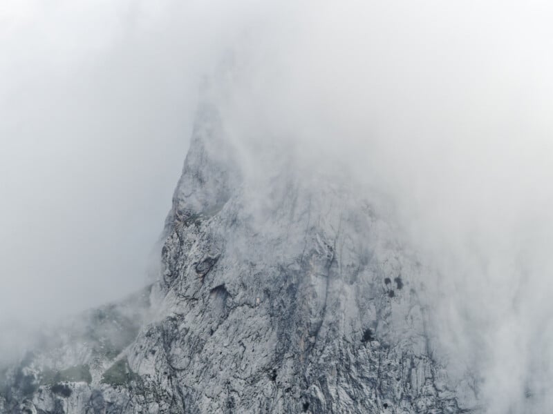 Un escarpado pico rocoso parcialmente envuelto por una espesa niebla blanca, cuyos escarpados acantilados y escasa vegetación apenas son visibles a través de la niebla.