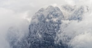 A rugged, rocky mountain peak partially obscured by thick, swirling clouds and mist, giving the scene a mysterious and dramatic atmosphere.