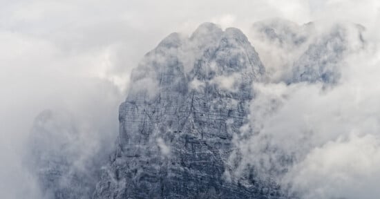 A rugged, rocky mountain peak partially obscured by thick, swirling clouds and mist, giving the scene a mysterious and dramatic atmosphere.