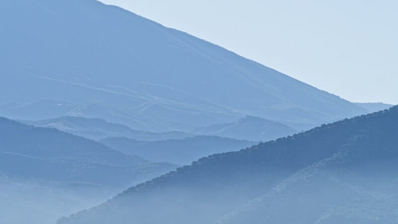 Las crestas montañosas azules se alejan gradualmente en la distancia y el valle se llena de una suave niebla o niebla, creando una escena pacífica y pacífica bajo el cielo pálido.