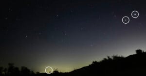 A night sky with many stars above a dark landscape; three white circles highlight specific stars in the sky and one on the horizon.