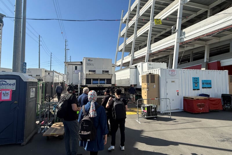 People stand in line outside temporary white tents and shipping containers next to a multi-level parking structure on a sunny day. Some are wearing backpacks; portable toilets and barriers are visible nearby.