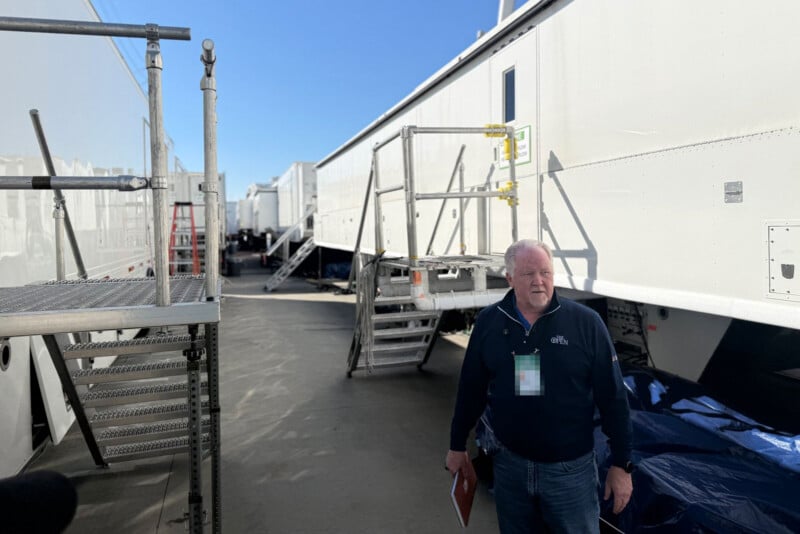 A man stands between two large white trailers with metal stairs, holding a folder. It’s sunny, and the sky is clear and blue. Industrial equipment and scaffolding are visible in the background.