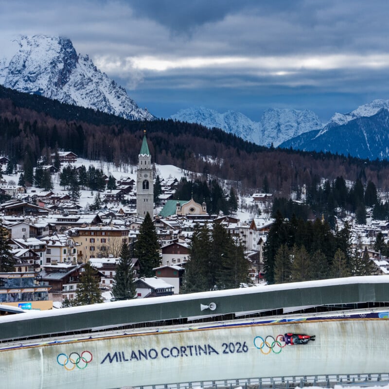 A bobsled races down an icy track marked "Milano Cortina 2026" with the Olympic rings, set against a snowy mountain village and majestic peaks in the background.