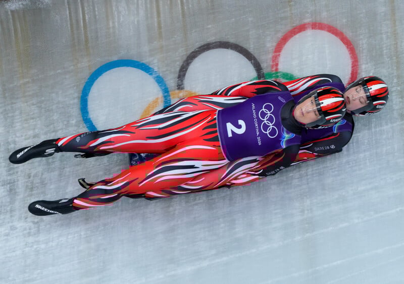 Two athletes in matching flame-patterned suits and helmets compete in a doubles luge event on an icy track with Olympic rings visible in the background.