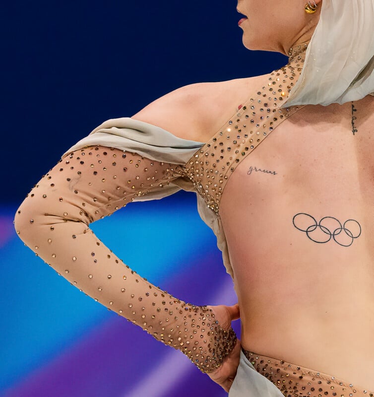 A figure skater in a sparkling, beige costume with sheer sleeves performs on ice. Visible are a tattoo of the Olympic rings and the word "grace" on their back, with a colorful blurred arena background.