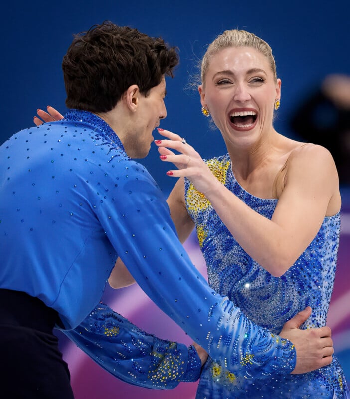 A smiling figure skating pair in sparkling blue costumes celebrate on the ice, the woman laughing joyfully with her arm raised as the man holds her waist.