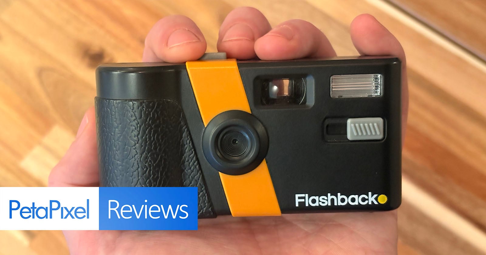 A hand holds a black and yellow point-and-shoot camera labeled "Flashback" over a wooden surface. "PetaPixel Reviews" text is visible in the lower left corner.