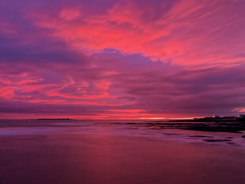 A vibrant sunset over the ocean with dramatic pink, purple, and orange clouds reflecting on calm water, and a dark silhouette of land on the horizon.