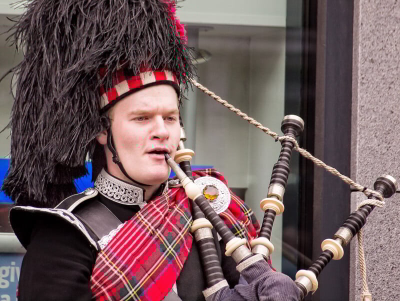 A man in traditional Scottish dress, including a feathered hat and red tartan sash, plays the bagpipes outdoors.