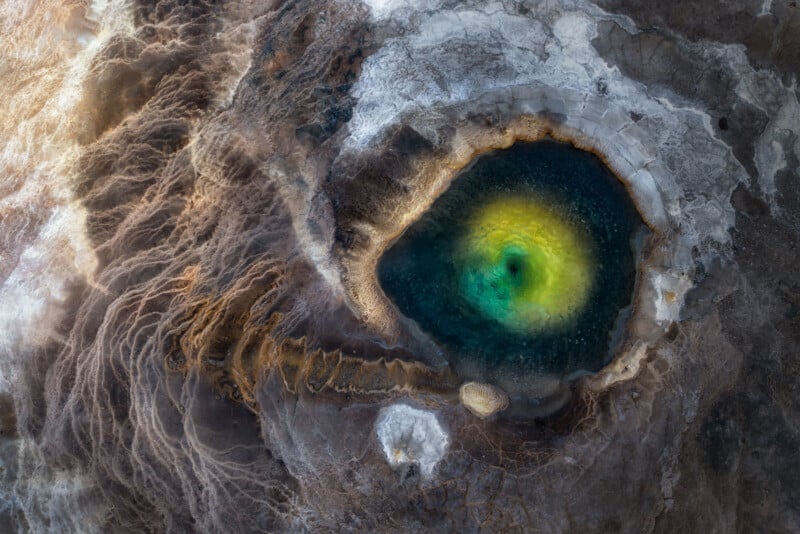 Aerial view of a vibrant geothermal spring with deep blue and green center, surrounded by yellow and white mineral deposits and textured brown earth formations.