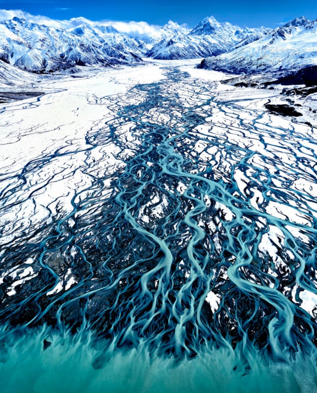 Aerial view of braided turquoise river channels winding through a snowy, mountainous landscape with rugged peaks in the background under a clear blue sky.
