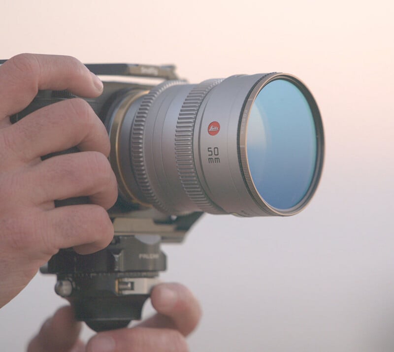 A close-up of hands holding a professional camera with a Leica 50mm lens, set against a soft, blurred background.