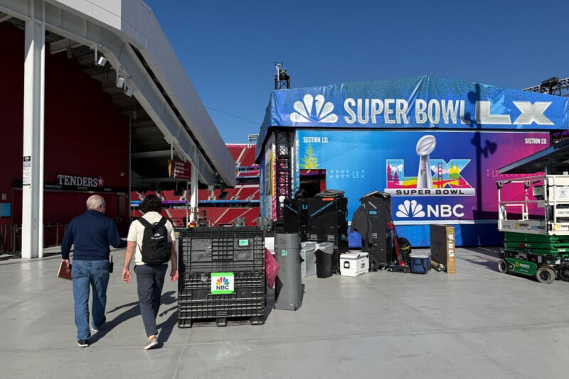 Two people walk toward a Super Bowl LX media setup at a stadium, passing equipment crates and NBC-branded items under a large blue Super Bowl LX sign.