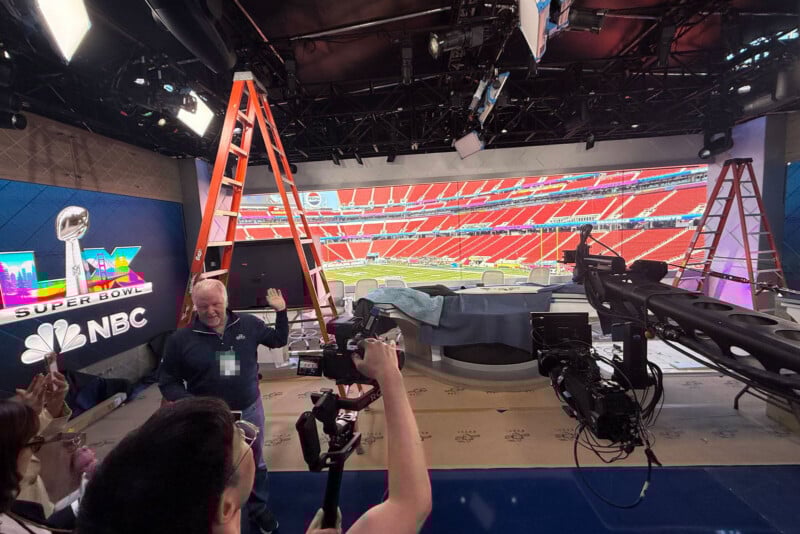 TV studio setup for NBC’s Super Bowl coverage, with cameras, two ladders, crew members, and a large screen showing an empty football stadium with red seats in the background.
