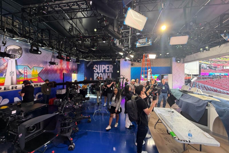 A busy TV studio with crew members, cameras, and equipment preparing for a Super Bowl broadcast. Bright lights hang from the ceiling, and the Super Bowl logo is displayed on a backdrop. A stadium is visible through the window.