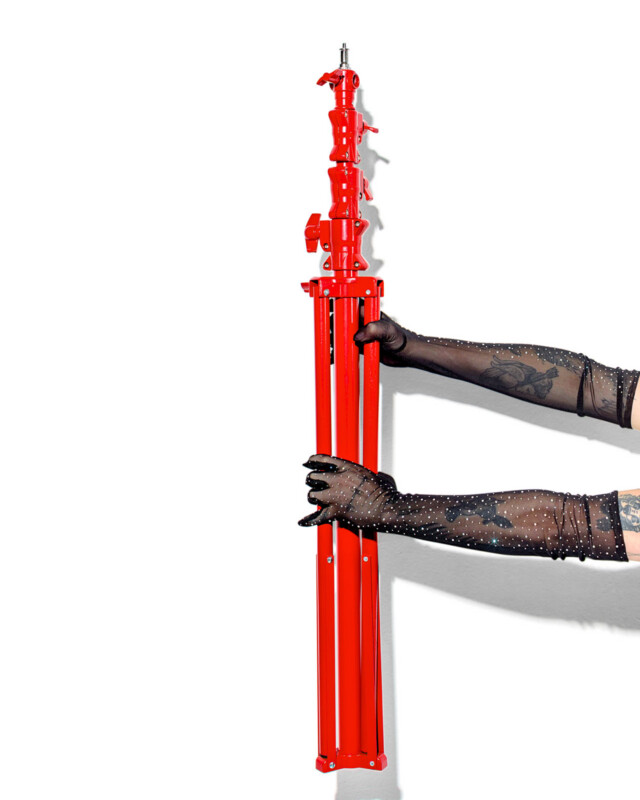 A person wearing black, sheer, beaded gloves holds a folded bright red tripod stand against a white background. Only the person's arms are visible, showing tattooed skin.