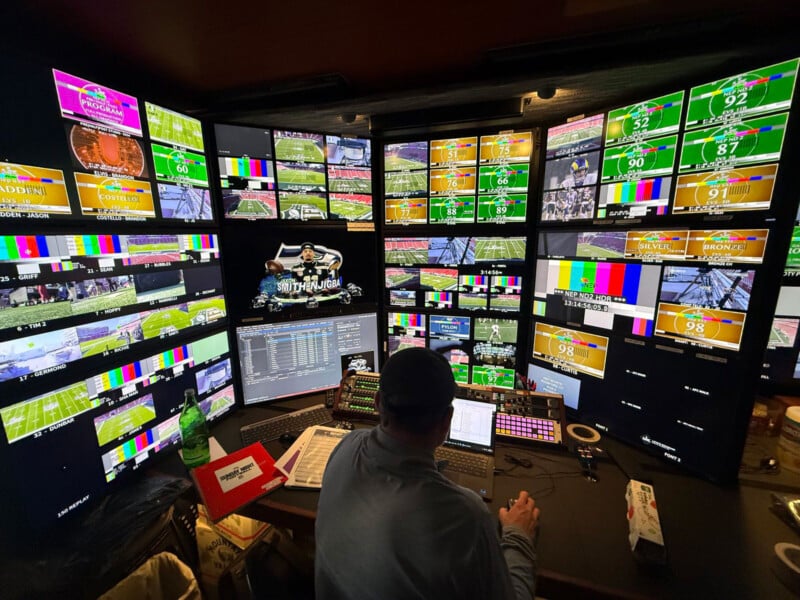 A person sits at a control panel in front of dozens of monitors displaying sports broadcasts, color bars, and camera feeds in a busy video production control room.