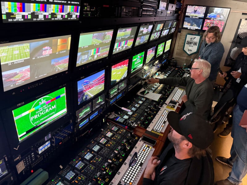 Several people work inside a broadcast control room, monitoring multiple screens displaying football game footage and graphics. The room is filled with equipment, control panels, and monitors.