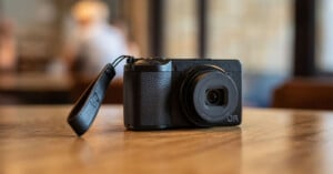 A compact black digital camera with a wrist strap rests on a wooden table in a softly lit indoor setting, with a blurred background of people and windows.