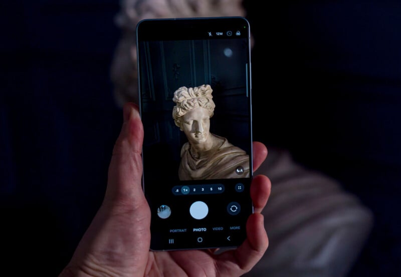 A hand holds a smartphone, capturing a photo of a classical marble bust. The phone screen shows the bust clearly, while the background is blurred.