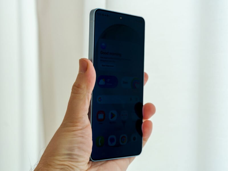 A hand holds a smartphone with a dark, reflective screen, showing faint app icons and text. The background is bright and out of focus, emphasizing the phone.