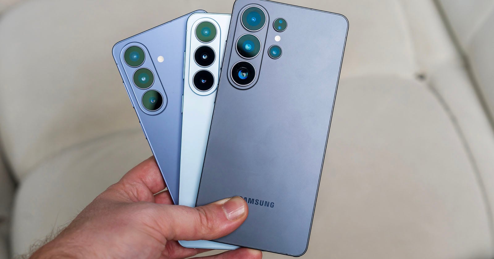 A hand holding three Samsung smartphones of different sizes and colors, showing the backs of the phones with their camera lenses prominently visible.