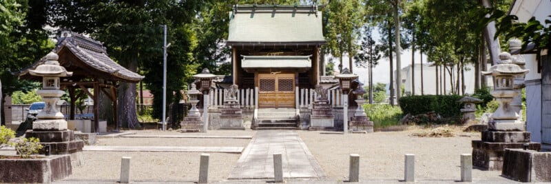 A traditional Japanese shrine with a wooden building, stone lanterns, and a tiled roof gate, surrounded by trees and a gravel courtyard under a clear sky.