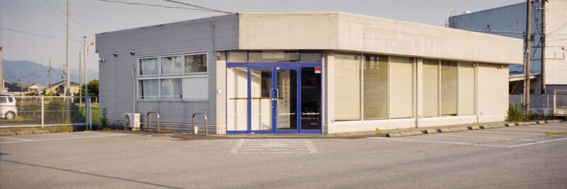 A small, rectangular, single-story building with large windows and a blue-framed glass door sits empty beside an open, sunlit parking lot lined with white markings. There are no cars or people present.