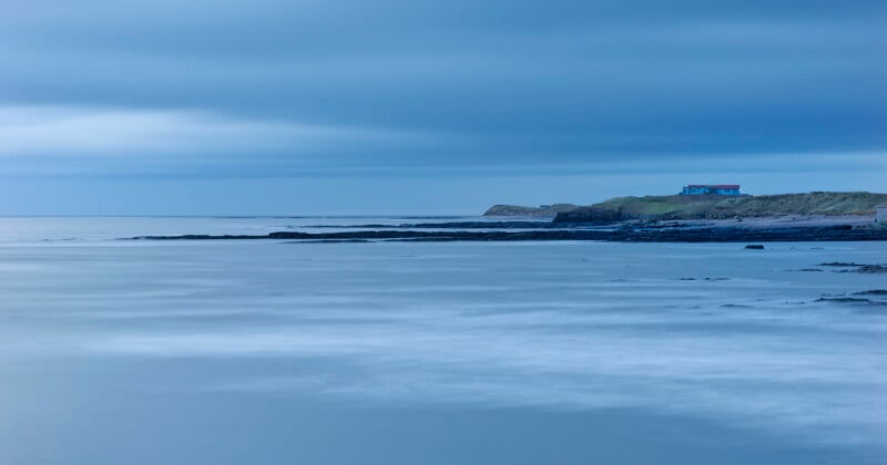 A calm, blue-toned seascape with smooth water, rocky shoreline, and a small house on a grassy cliff under a cloudy sky. The scene appears peaceful and serene.