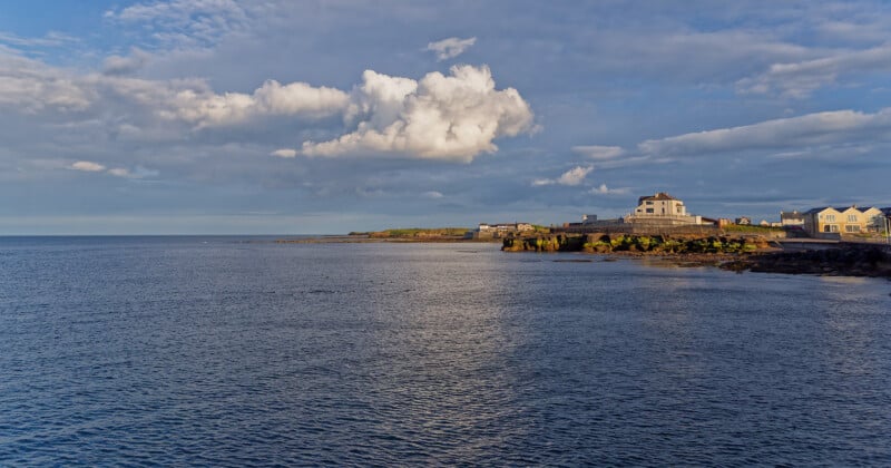 A calm coastal scene with blue water, a rocky shoreline, houses near the edge, and a partly cloudy sky. Sunlight illuminates the buildings and casts reflections on the sea.