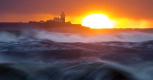 Waves crash in the foreground as the sun sets behind a distant lighthouse and buildings on a dark coastline, casting a dramatic orange and yellow glow across the sky and water.