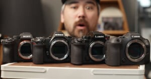 A surprised man stands behind four mirrorless camera bodies—Sony, Nikon, Panasonic Lumix, and Canon—displayed side by side on a table.