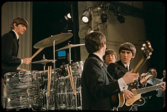 Four musicians in suits perform on stage. One plays drums while the others play guitar and bass. Bright stage lights shine overhead, and an audience watches in the background.