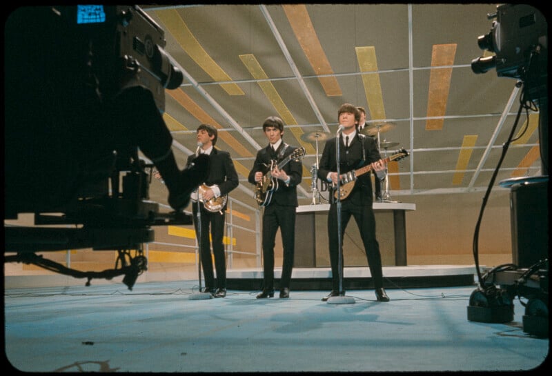 Three musicians in suits perform on a brightly lit stage with geometric patterns, holding guitars, as a camera films them; a drummer sits behind them at a drum kit.