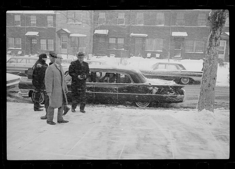 Four men stand beside a black car parked on a snowy street during snowfall. One man is getting into the back seat, while others wear coats and hats. Snow covers the ground, cars, and buildings in the background.