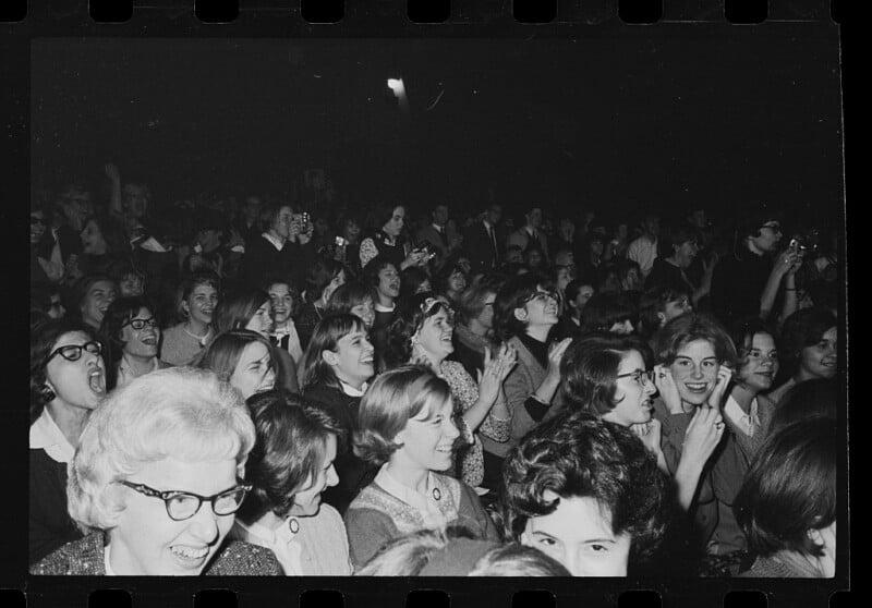 A large crowd of mostly young women, many smiling and cheering, sit closely together in a dark auditorium, reacting excitedly to something happening off-camera.