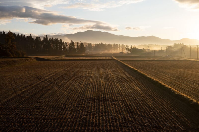 A wide, plowed field is bathed in warm sunset light, with trees lining the horizon and misty mountains in the distance under a partly cloudy sky.